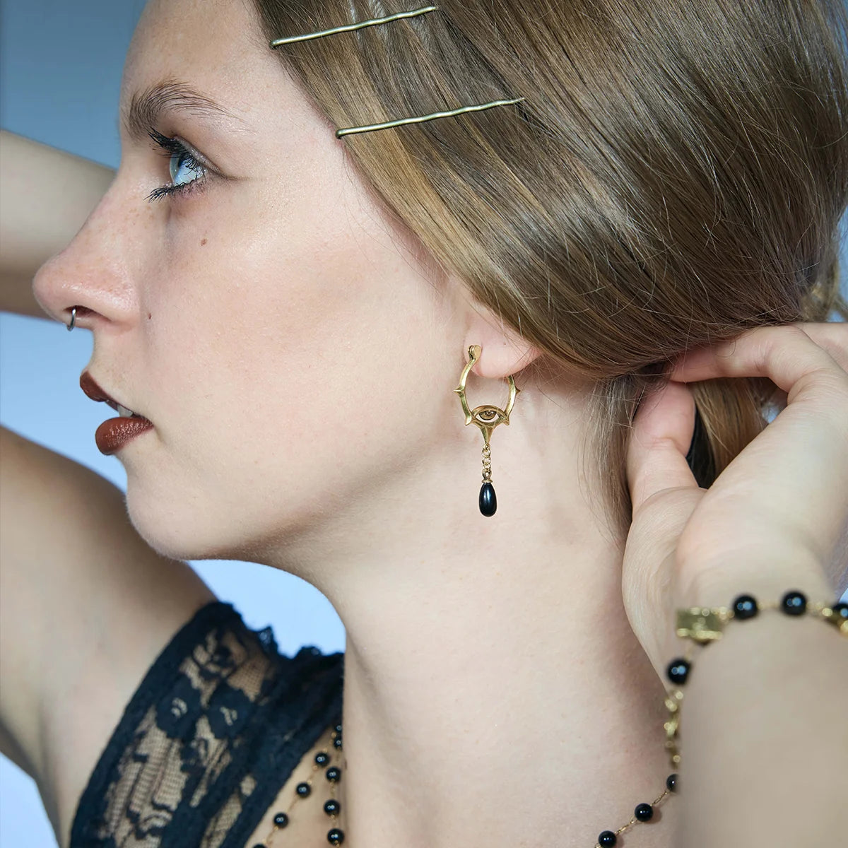 Side portrait showing a gold-tone Thorned Gaze Hoop Earring hanging below the lobe with an engraved eye motif and black teardrop accent.