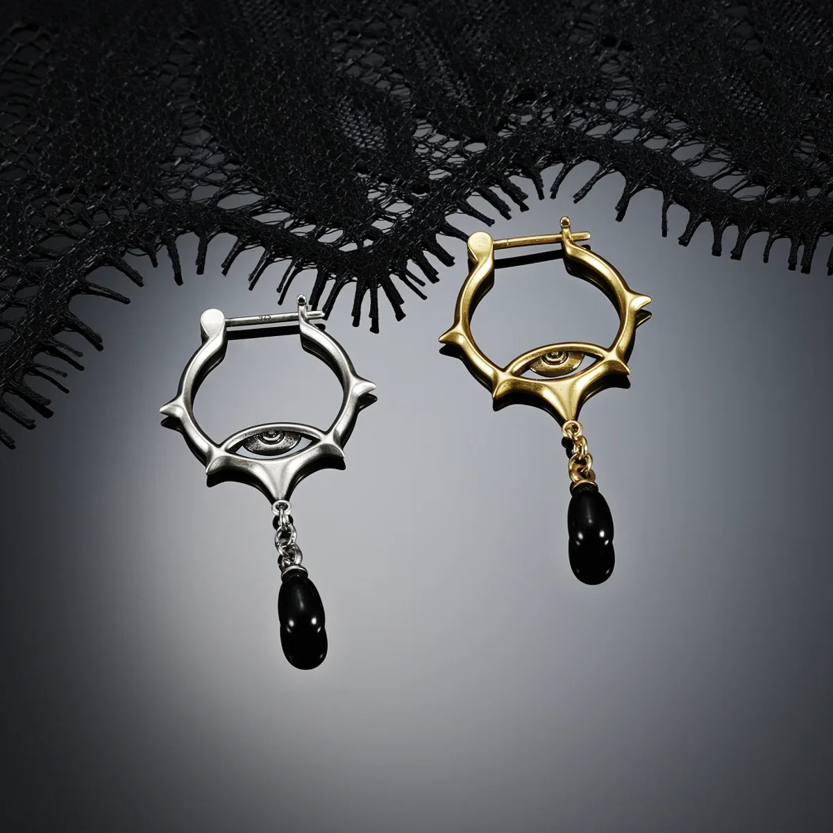 Antiqued silver- and gold-tone Thorned Gaze Hoop Earrings displayed together against black lace on a dark reflective surface.