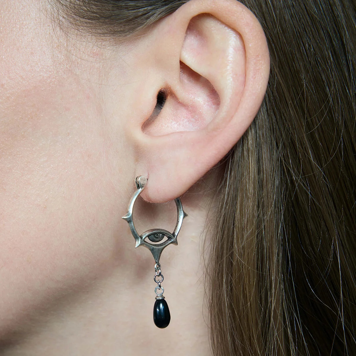 Close-up of an antiqued silver-tone Thorned Gaze Hoop Earring worn from the side, featuring an engraved eye motif and a suspended black teardrop accent.