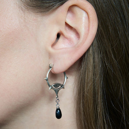 Close-up of an antiqued silver-tone Thorned Gaze Hoop Earring worn from the side, featuring an engraved eye motif and a suspended black teardrop accent.