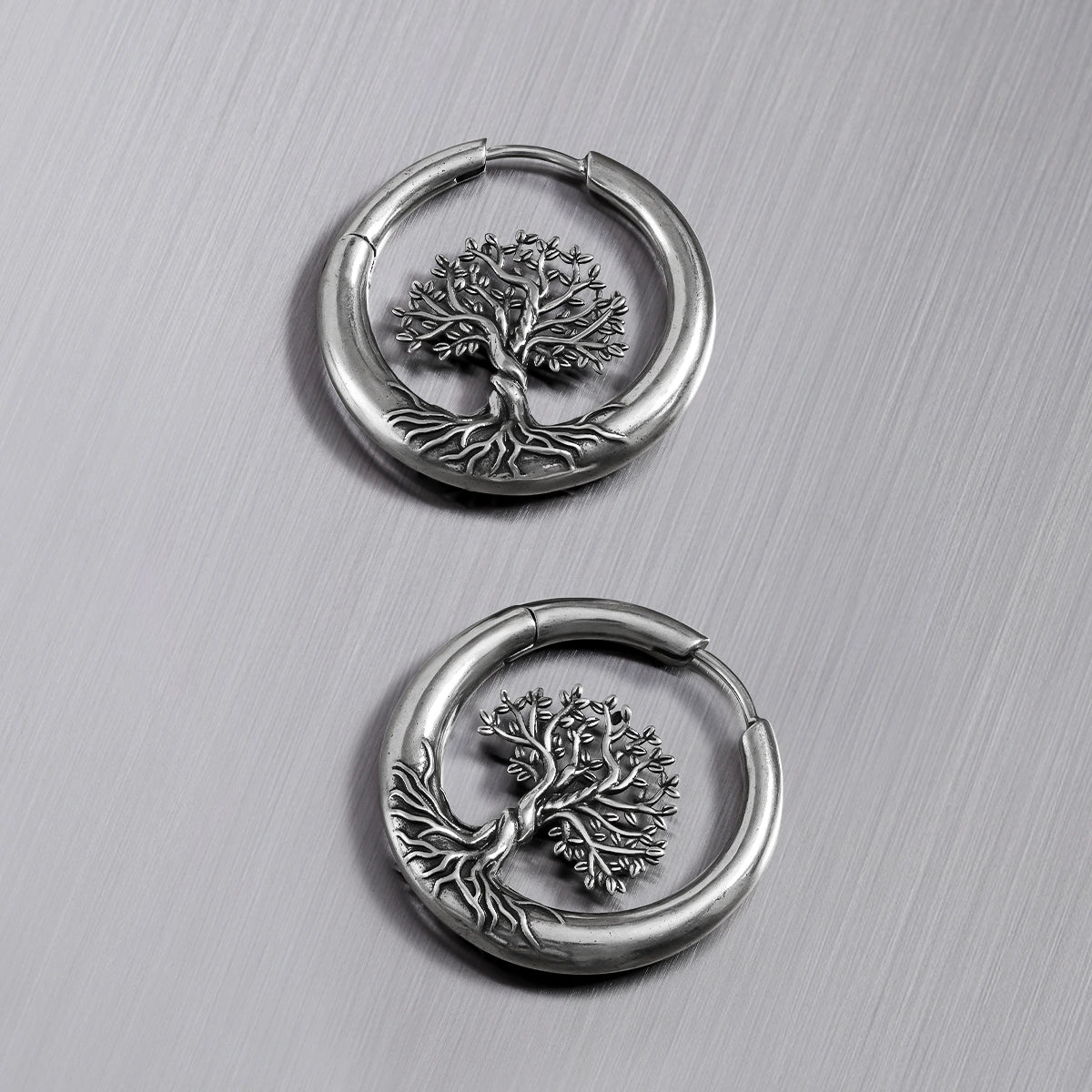 Pair of silver-tone tree of life hoop earrings laid flat on a metallic surface