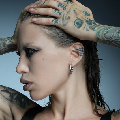 Model with tattooed hands touching the head wears a small silver-tone hoop earring with horn details