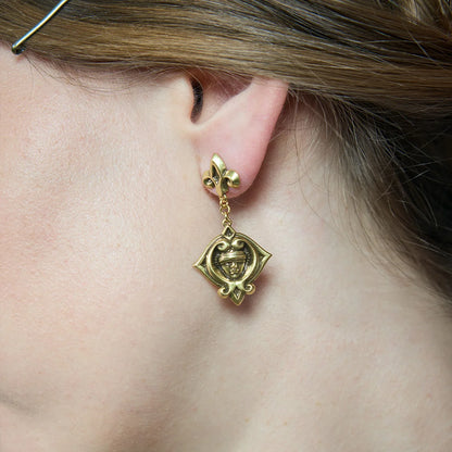 Rear close-up of a gold-tone Veiled Seer Drop Earring worn below the lobe, showing the suspended medallion and fleur-de-lis stud.