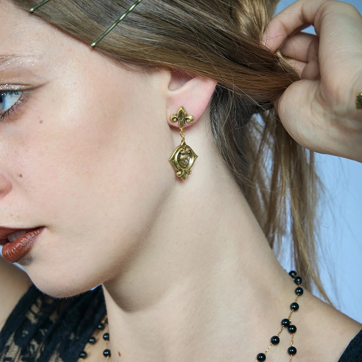 Side portrait of a gold-tone Veiled Seer Drop Earring with the hair lifted to reveal the blindfolded face motif.