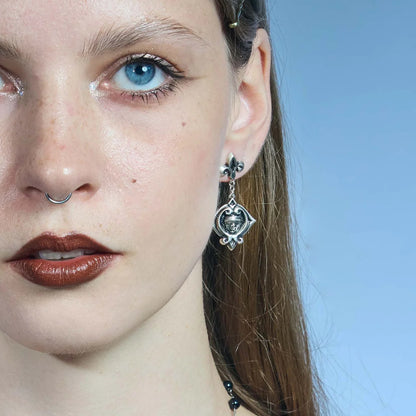 Front close-up of an antiqued silver-tone Veiled Seer Drop Earring with a blindfolded face motif and pointed eye-shaped frame.