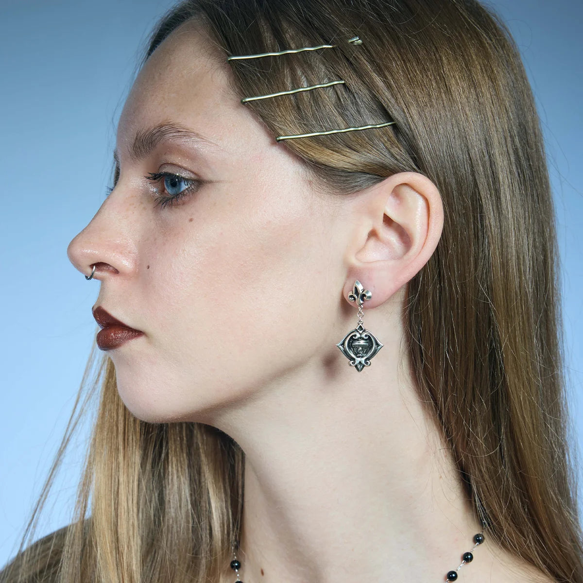 Side portrait showing an antiqued silver-tone Veiled Seer Drop Earring suspended below a fleur-de-lis stud.