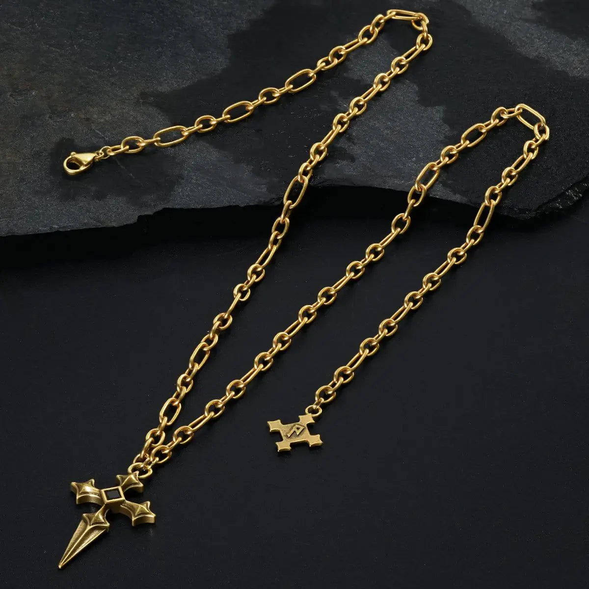 Gold Vesper Blade Necklace laid across black textured fabric with the blade-shaped cross pendant in view.