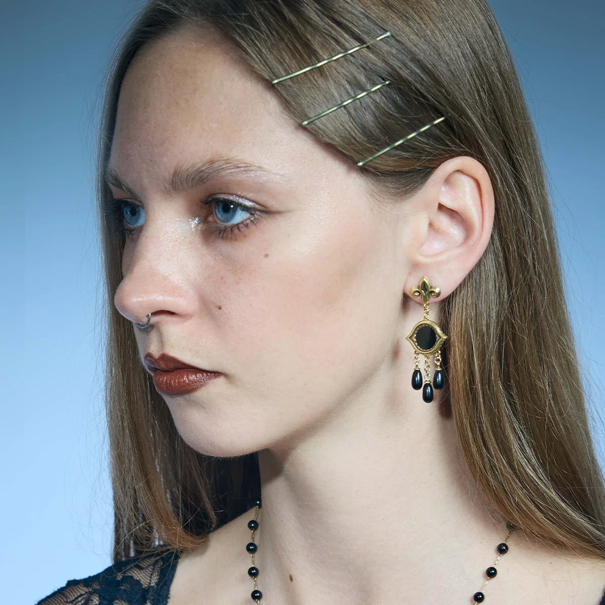 Side portrait showing a gold-tone Votive Mirror Earring hanging below the lobe with three suspended black drops.
