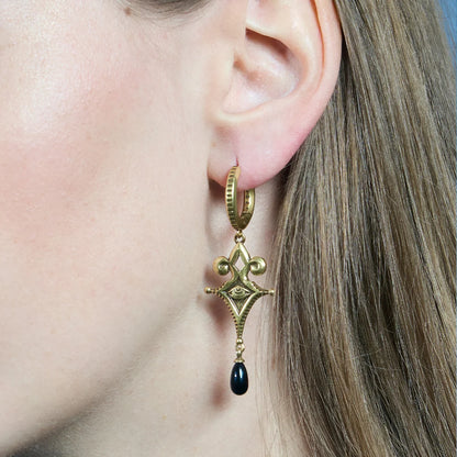 Close-up of a gold-tone Watcher’s Tear Earring worn from the side, featuring an engraved eye motif and a suspended black teardrop accent.