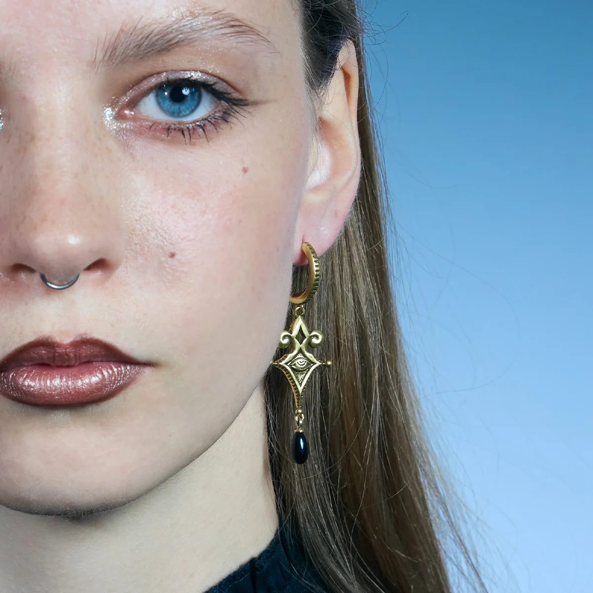 Front close-up of a gold-tone Watcher’s Tear Earring, showing the pointed ornamental frame and narrow black teardrop accent.