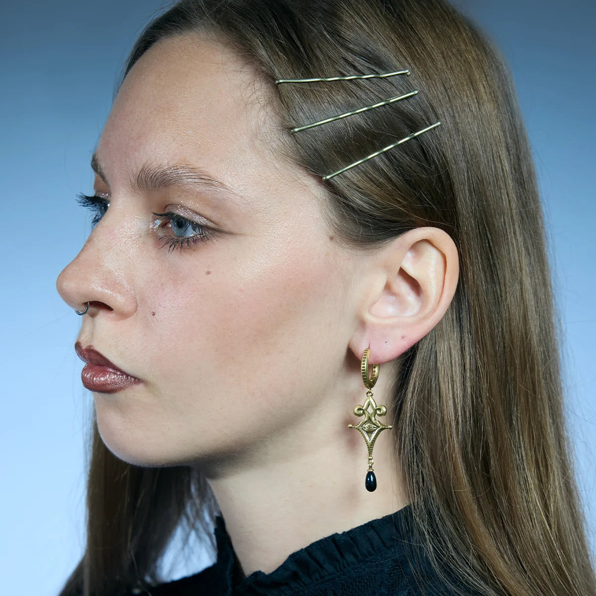 Side portrait showing a gold-tone Watcher’s Tear Earring hanging below the lobe with an engraved eye motif and black teardrop accent.