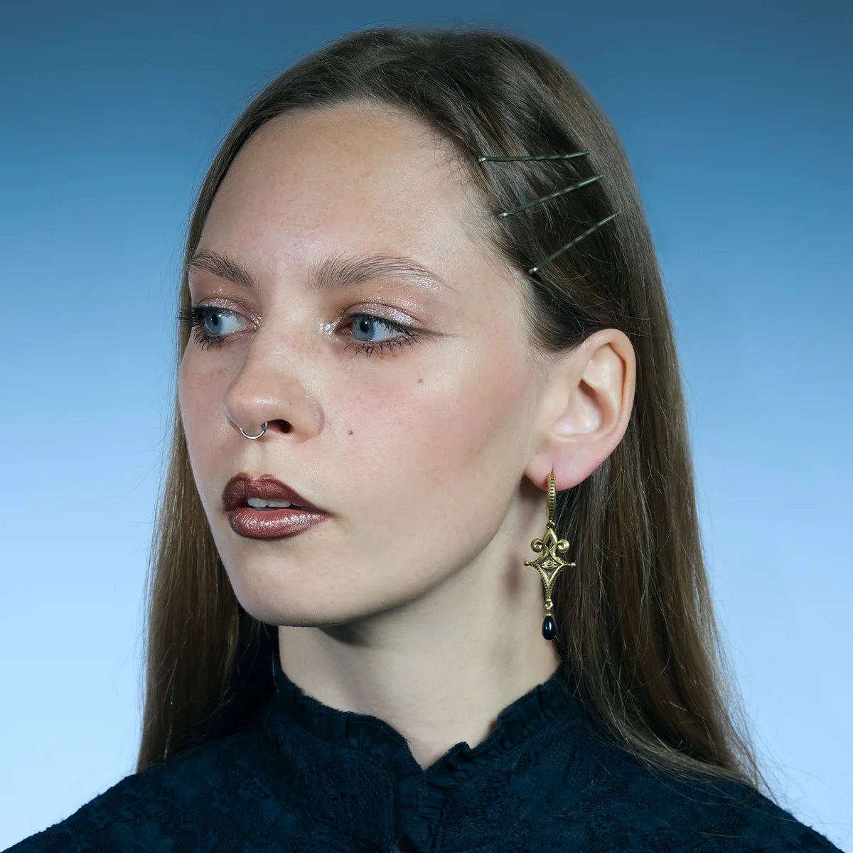 Three-quarter portrait showing gold-tone Watcher’s Tear Earrings with pointed ornamental frames and suspended black teardrop accents.