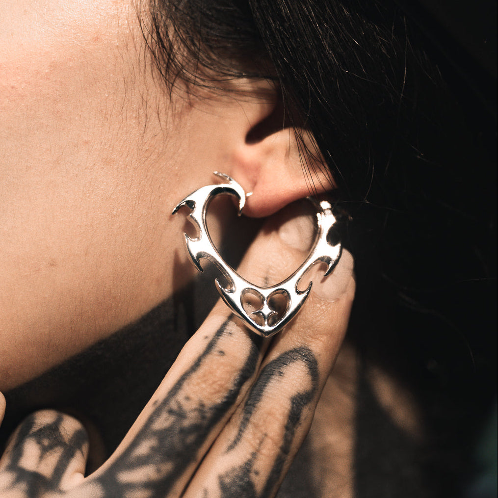 Silver-tone wild heart earring with jagged edges worn on ear, close-up with hand near tattooed skin