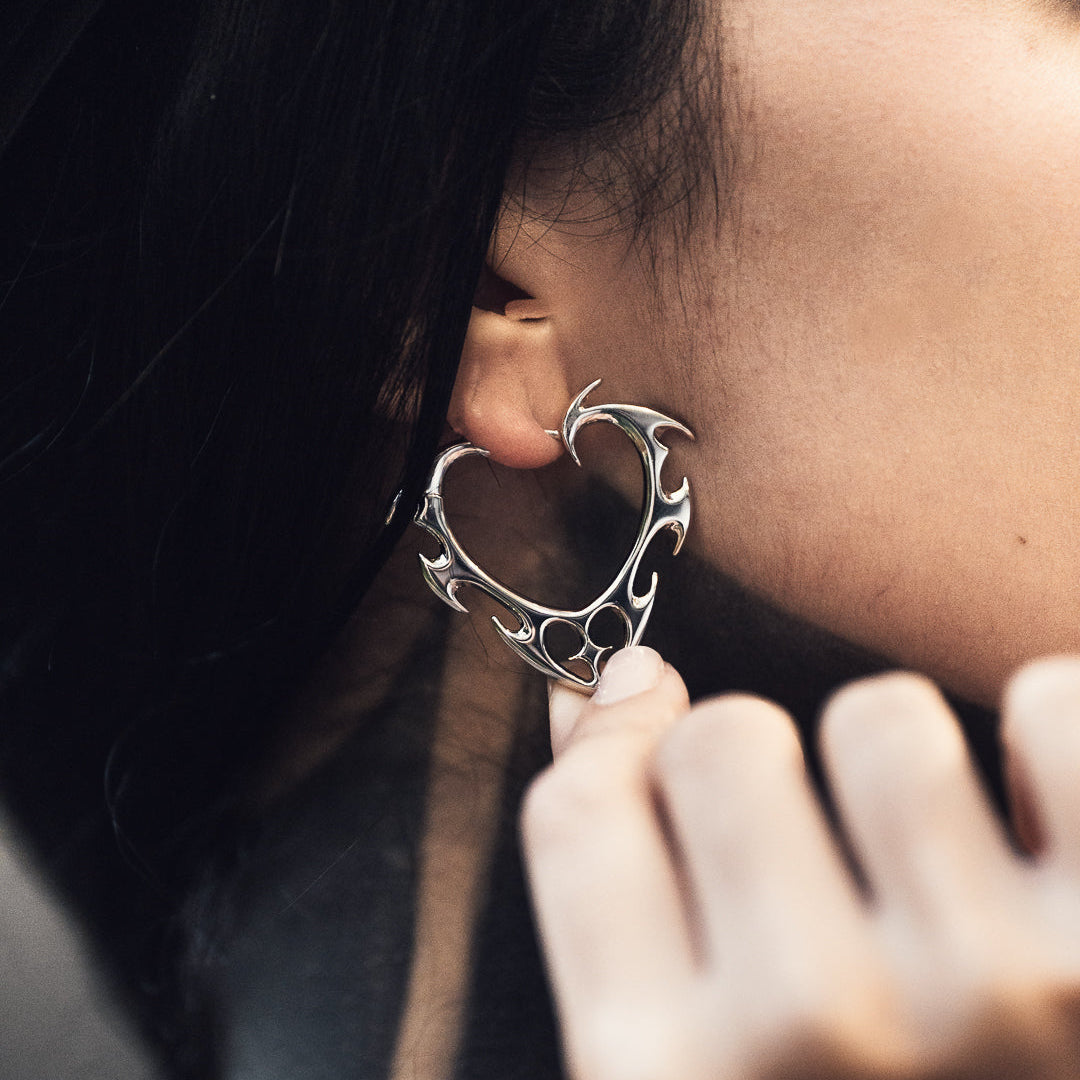 Close-up of silver-tone wild heart earring with thorn details worn on ear, fingers gently touching it