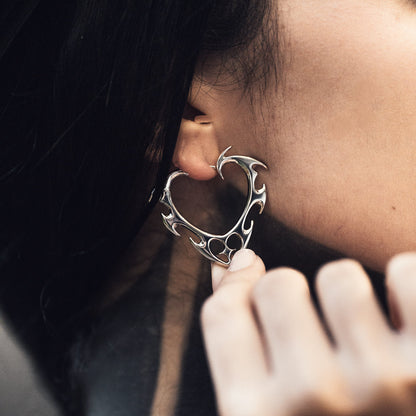 Close-up of silver-tone wild heart earring with thorn details worn on ear, fingers gently touching it