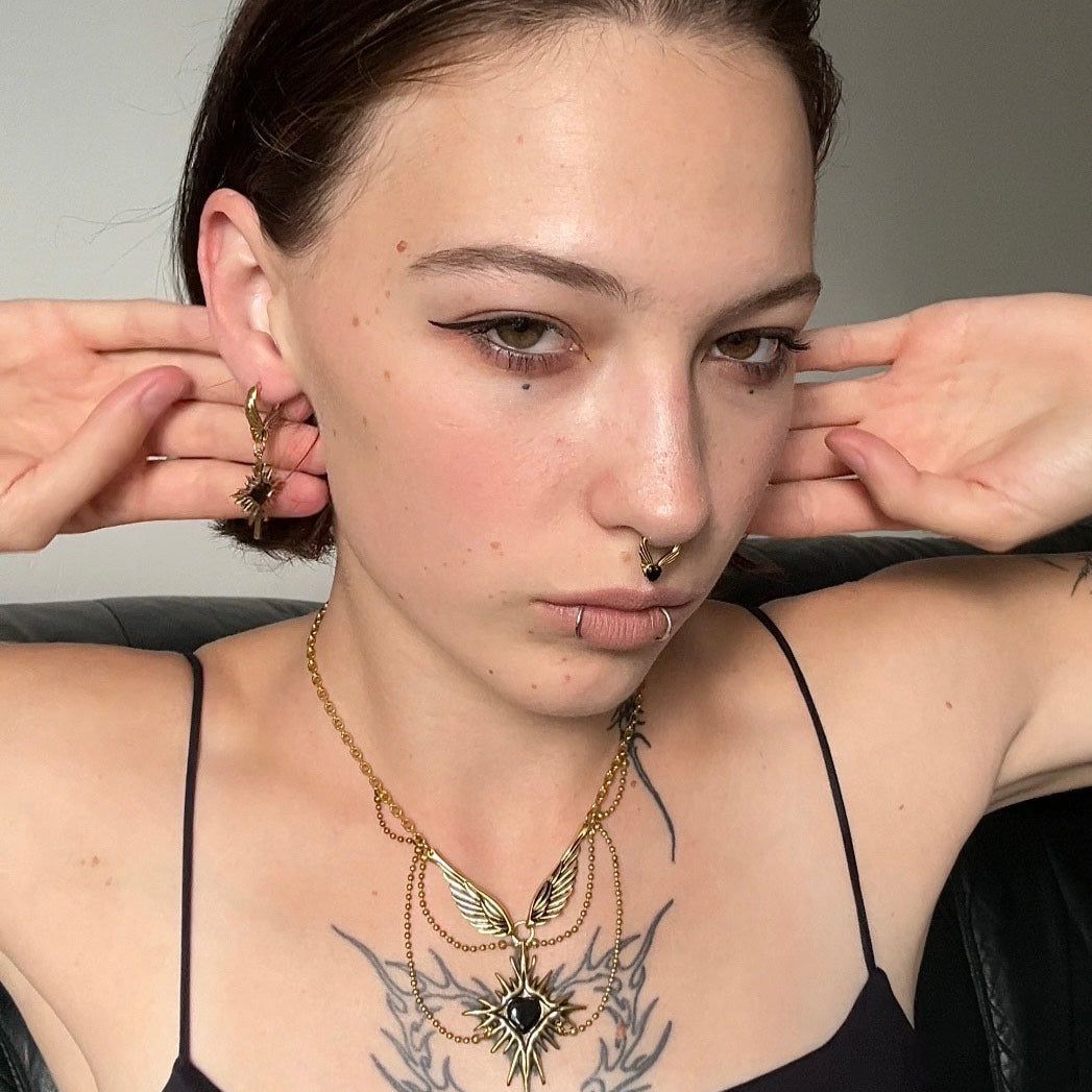 Winged Heartburst Earrings in 925 sterling silver, worn in a front portrait with raised arms, a chest tattoo, and a black cam