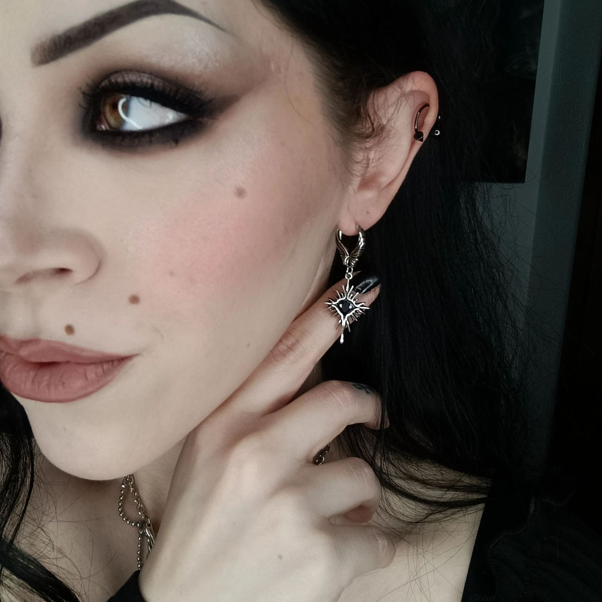 Winged Heartburst Earrings in 925 sterling silver, worn in a side portrait with smoky eye makeup and a hand resting at the ne