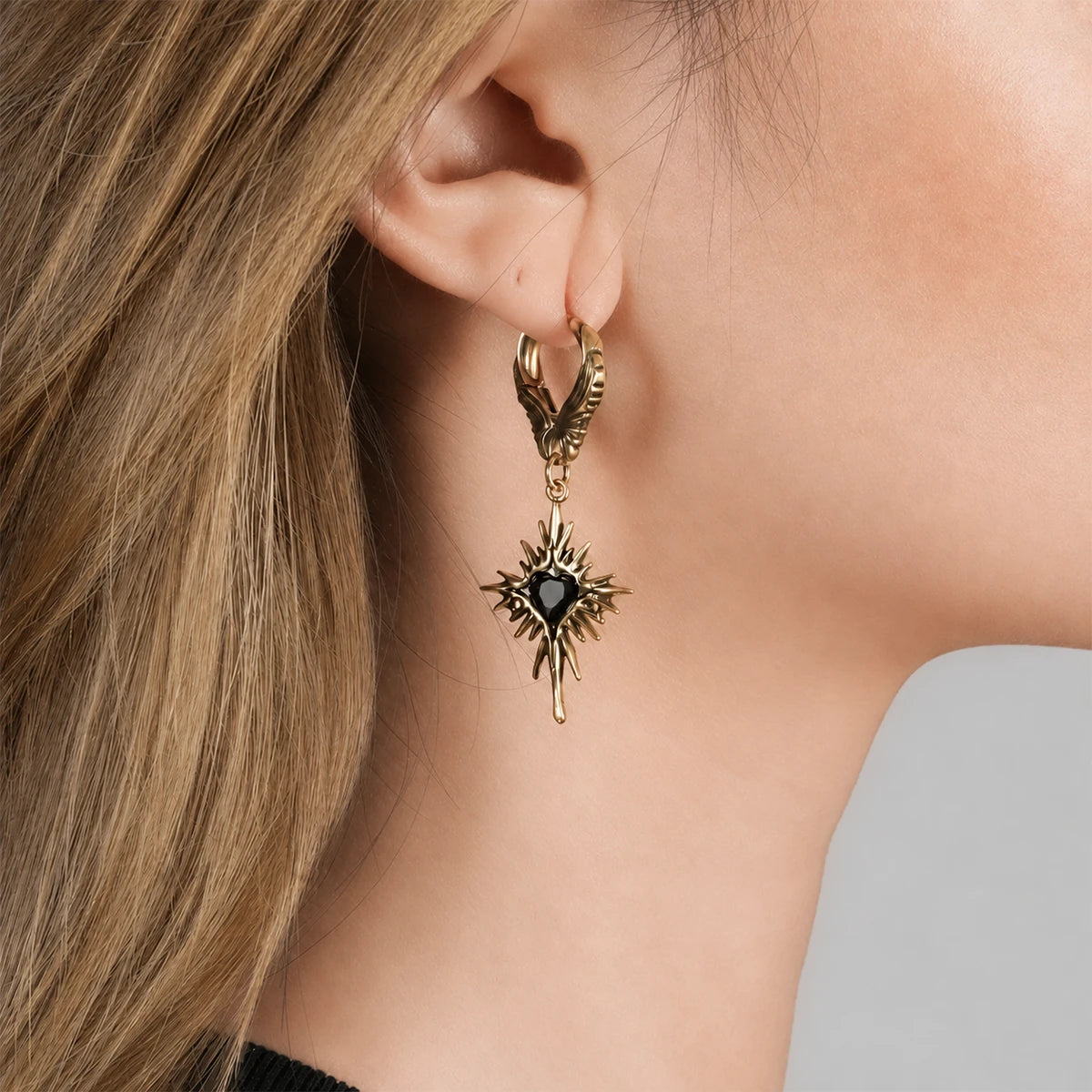 Gold-tone Winged Heartburst hoop earring worn on an earlobe, with a black heart and radiating spikes