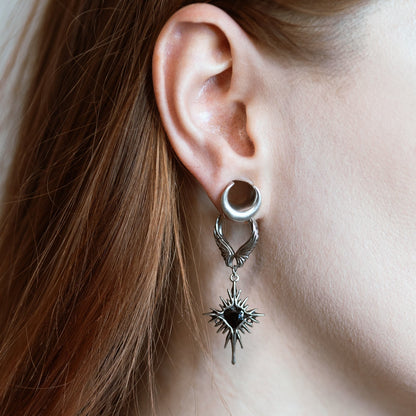 Winged hoop earring with a heart-centered starburst charm, worn below a silver-tone ear tunnel