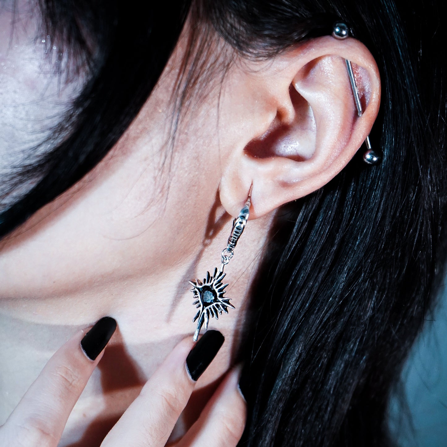 Silver-tone starburst earring with dark center hanging from a hoop, worn alongside an ear piercing